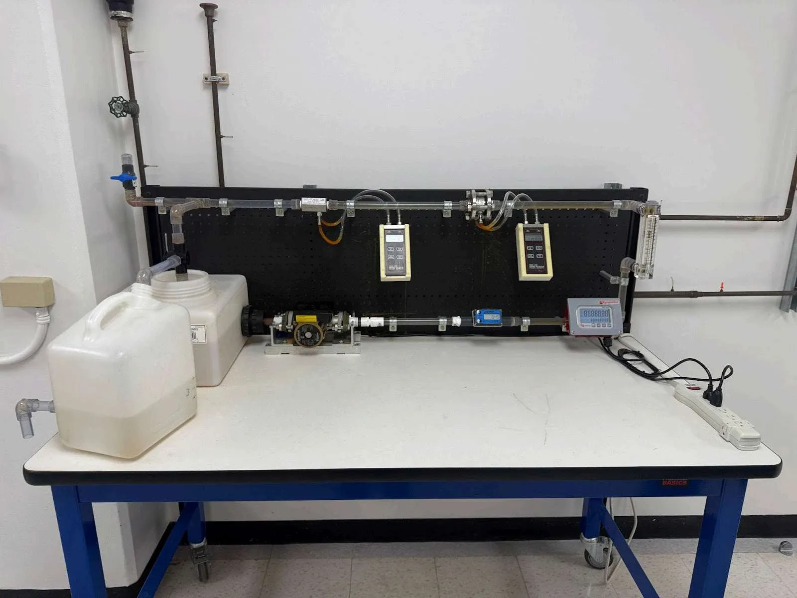 Photograph of the laboratory flow bench: a closed water loop on a bench frame with the five inline flow meters, gauges, and a reservoir jug