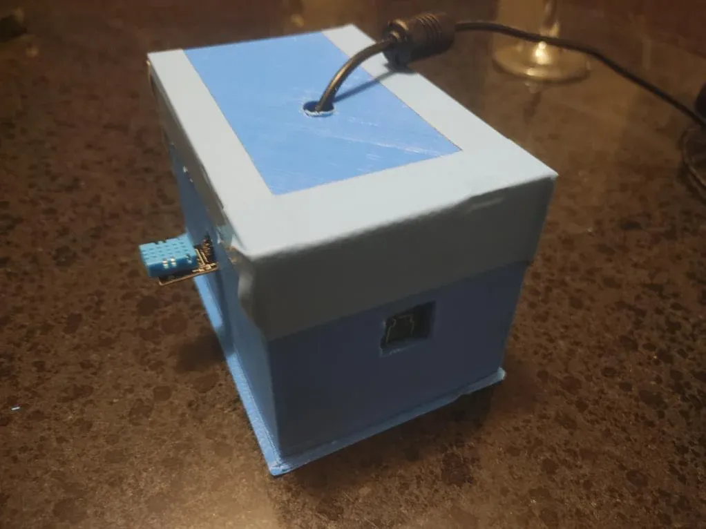 The final prototype: a light blue 3D-printed box on a countertop with a square camera window on the front, a sensor board on the side, and a cable exiting the top