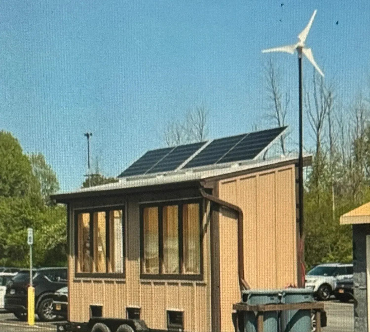 Side view of the finished trailer-mounted greenhouse in a parking lot: two solar panels on the sloped roof, large windows, and the 400 W wind turbine on a mast behind it