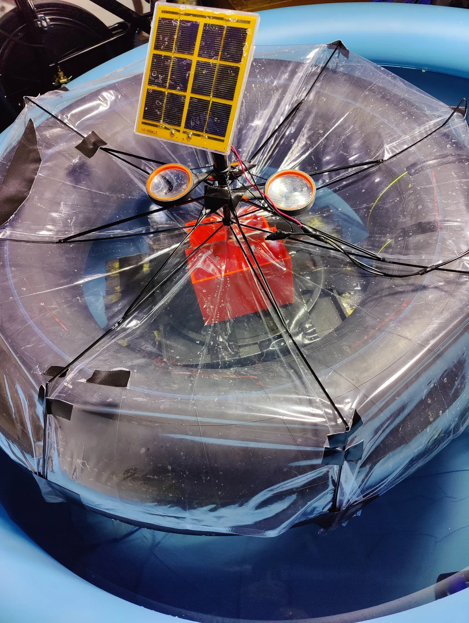 Floating desalination prototype in a test pool: an inverted clear umbrella frame with taped film panels, a small solar panel mounted at the center mast, and the red 3D-printed electrolysis chamber visible beneath the film