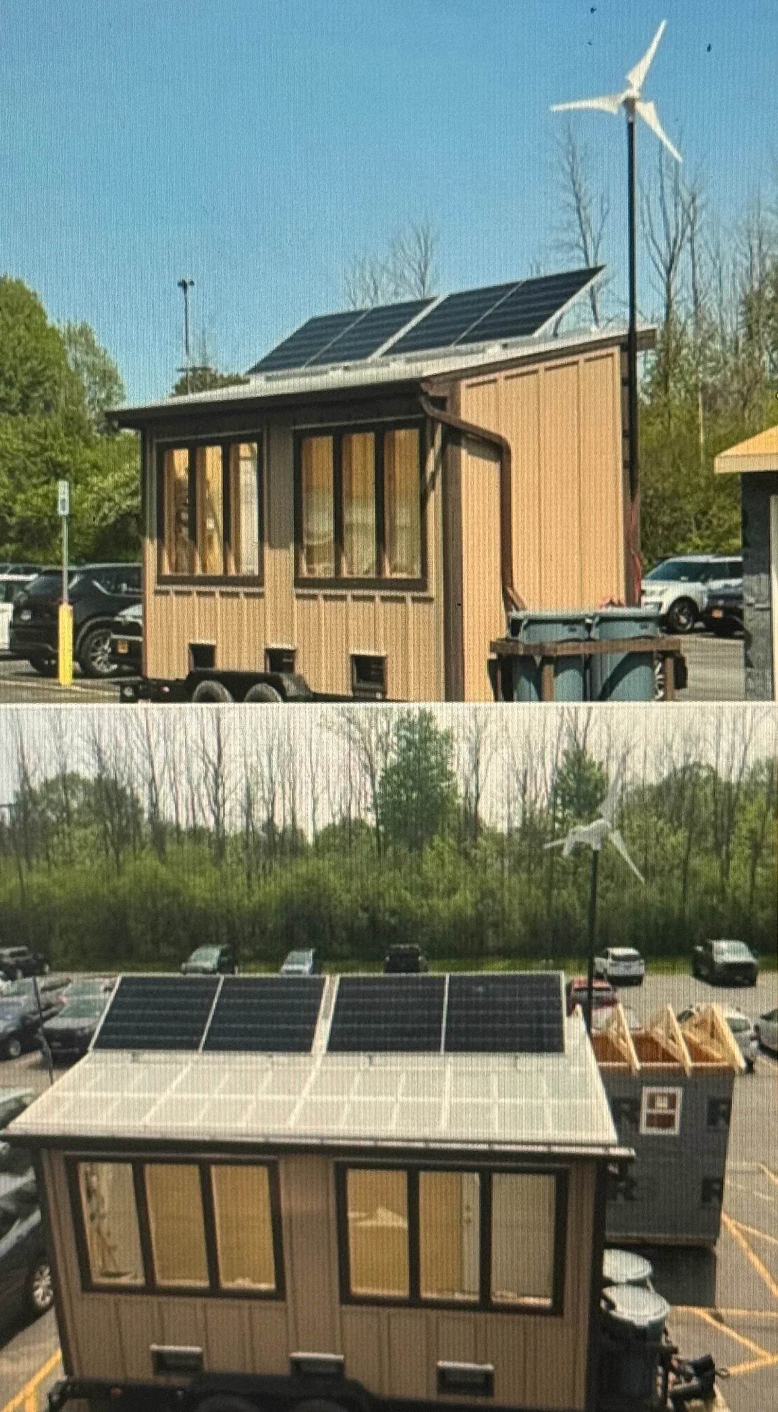 Completed trailer-mounted greenhouse in a parking lot with two rooftop solar panels on a sloped translucent roof and a wind turbine on a mast behind it