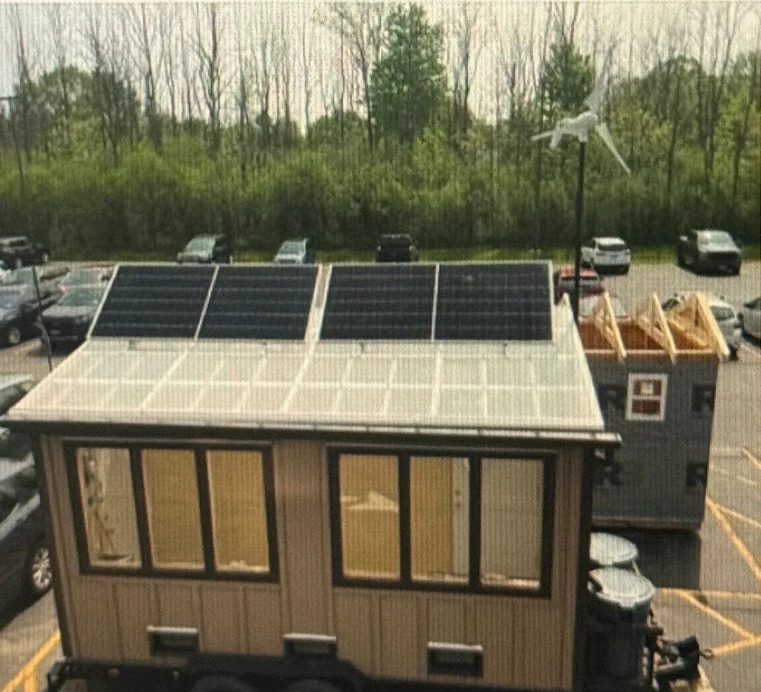 View from above of the finished greenhouse showing both solar panels mounted above the translucent roof panels, with the wind turbine on its mast and the parking lot behind