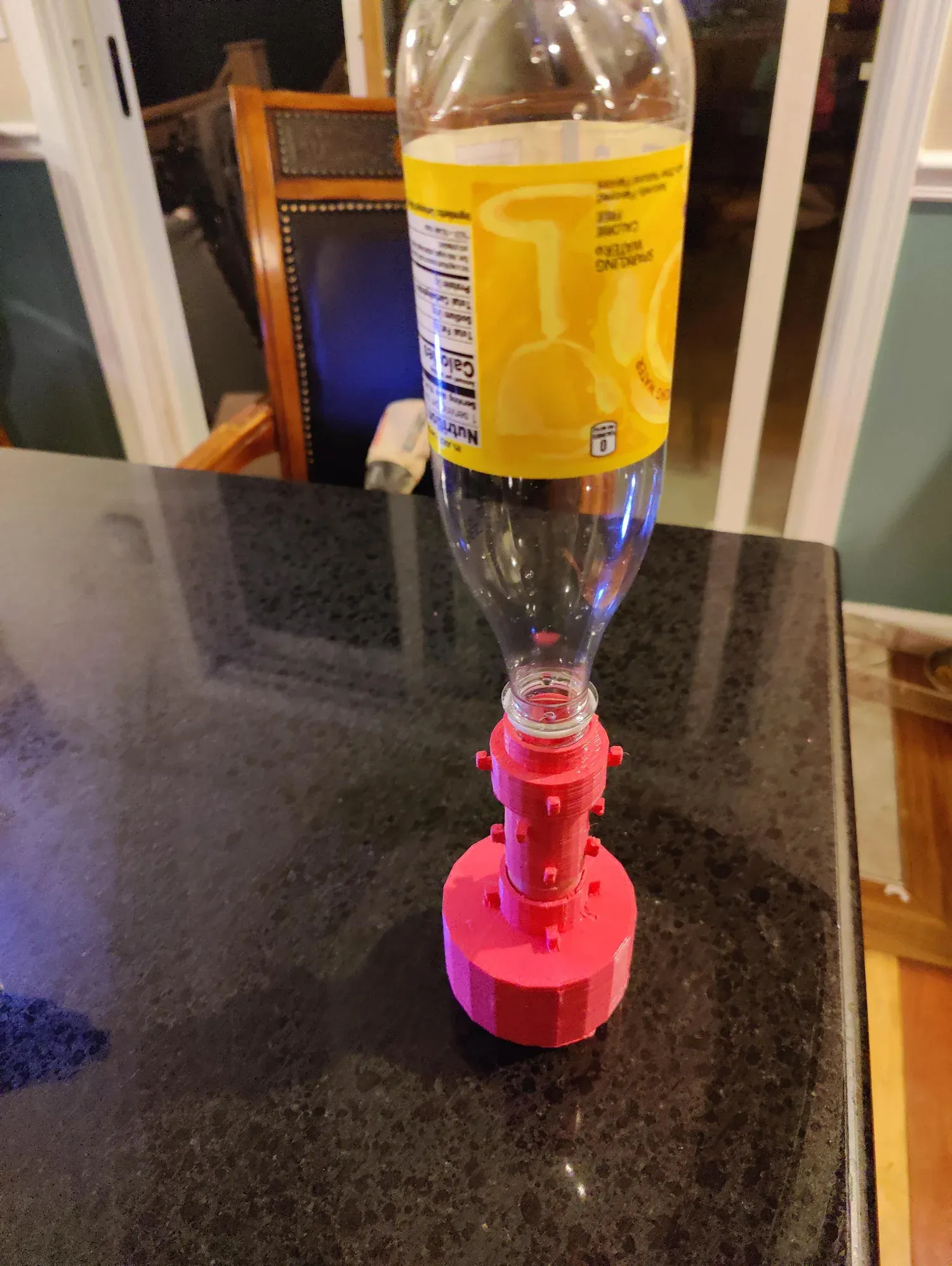 The red FDM 3D-printed electrolysis chamber on a countertop with an inverted clear plastic bottle seated in its collection port