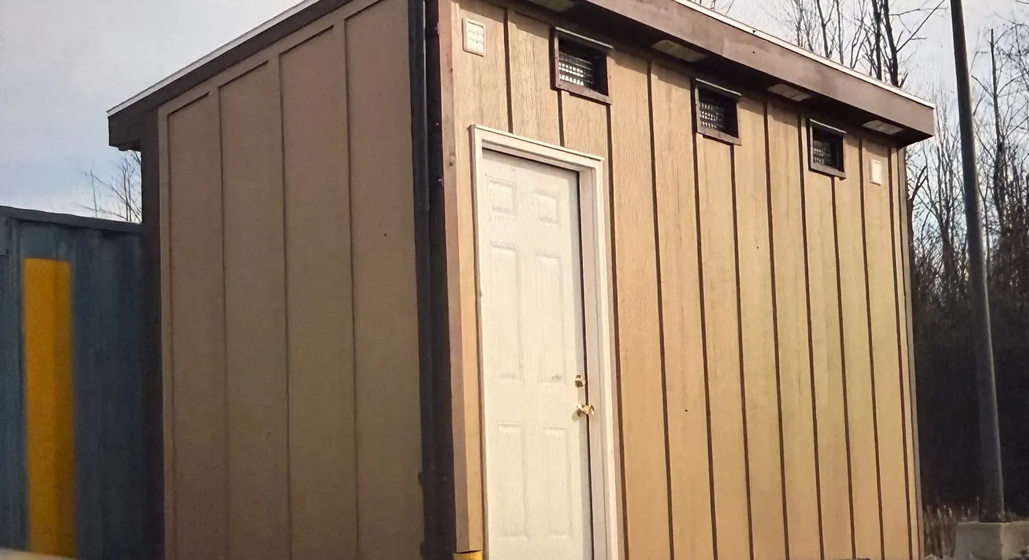 Exterior of the greenhouse during construction with tan board-and-batten siding, a white entry door, and small vent openings under the roofline