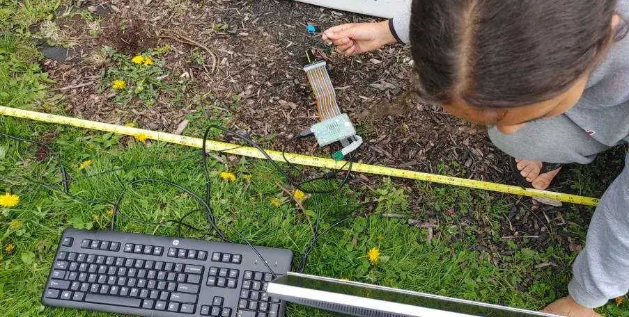 Testing outdoors: the device on the grass beside a tape measure laid out toward the fire, with a keyboard connected for reading the sensor output