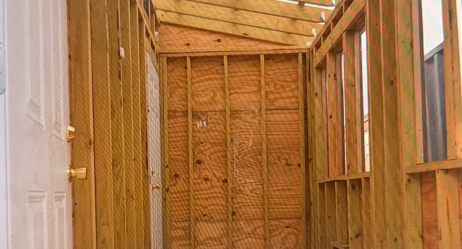 Interior of the greenhouse during construction showing exposed wood stud framing, roof rafters, and window openings