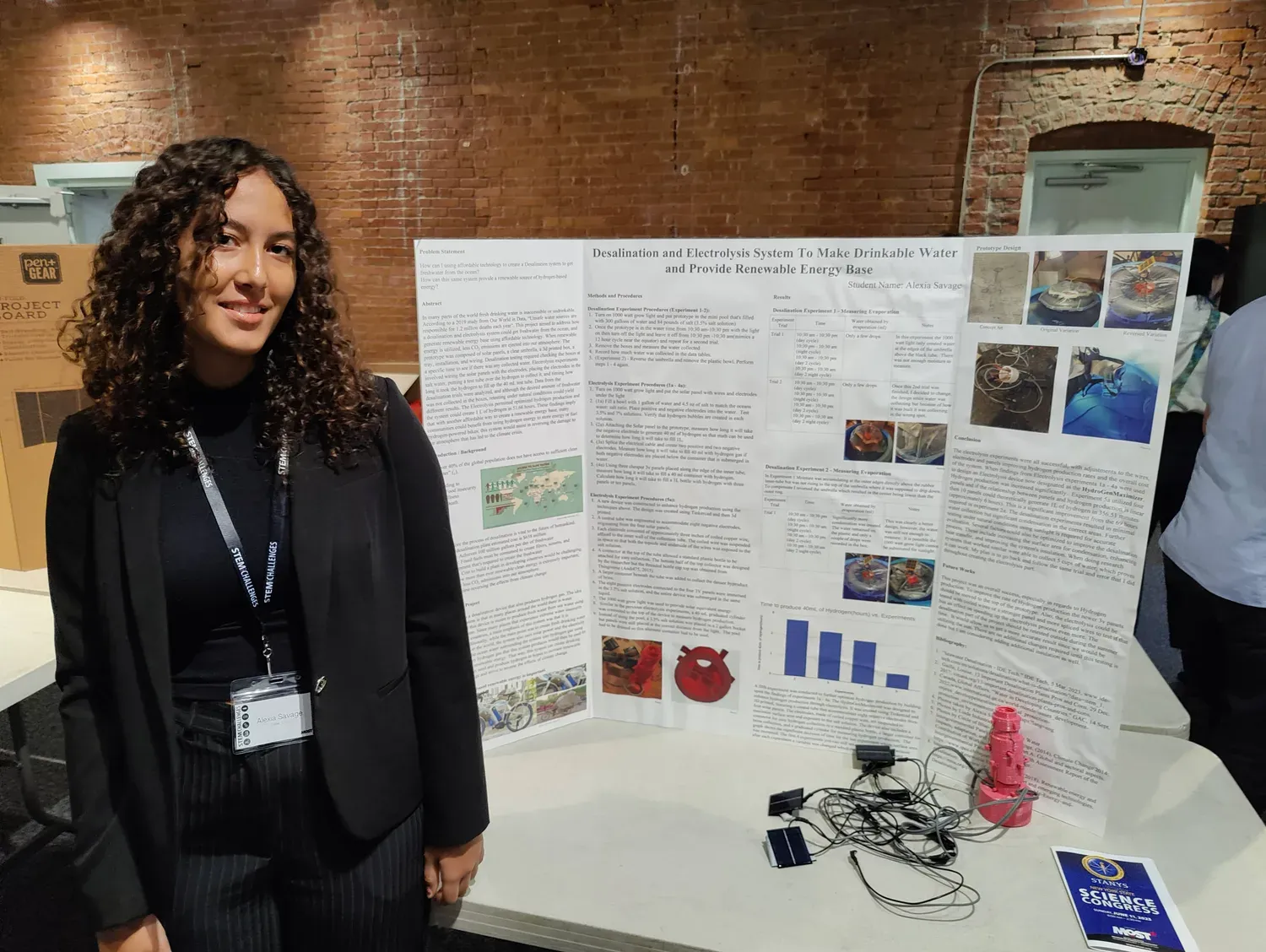 Alexia standing beside her research poster and hardware at the STANYS New York State Science Congress, with solar panels and the red electrolysis chamber on the table