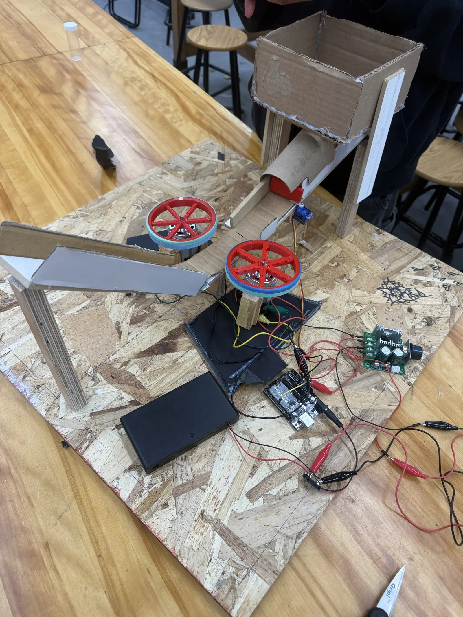 The built launcher on a bench: a cardboard reservoir on a wooden stand feeds a cardboard tube and ramp down to two red 3D-printed flywheels, with the Arduino, PWM controller, and battery pack wired on the OSB base