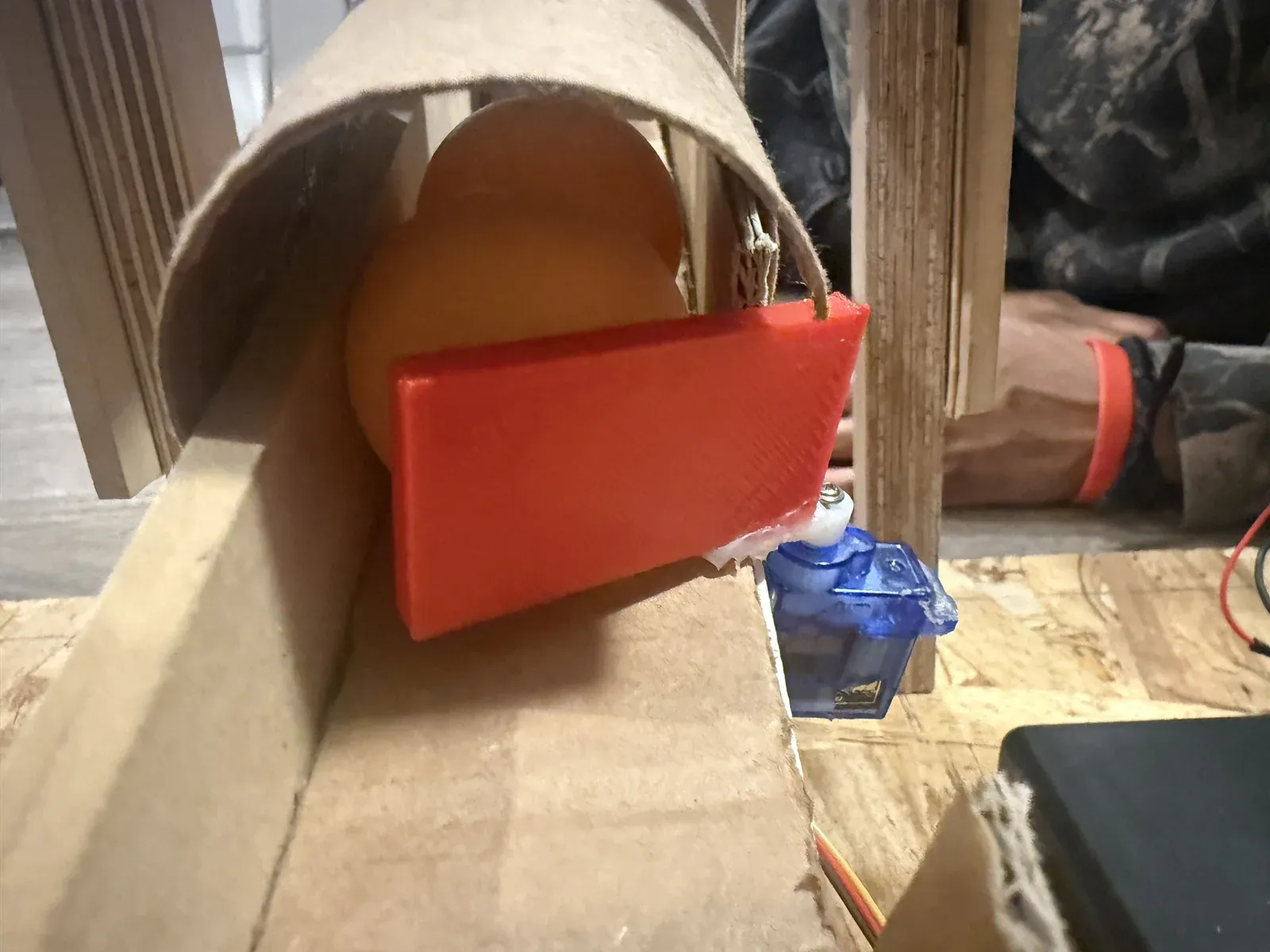 Close-up of the 3D-printed red door at the bottom of the cardboard tube holding back an orange ping-pong ball, with the small blue servo that opens it mounted beside the ramp